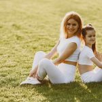 A mother and daughter sit back to back in matching activewear on a sunny grass field, enjoying a summer day together.